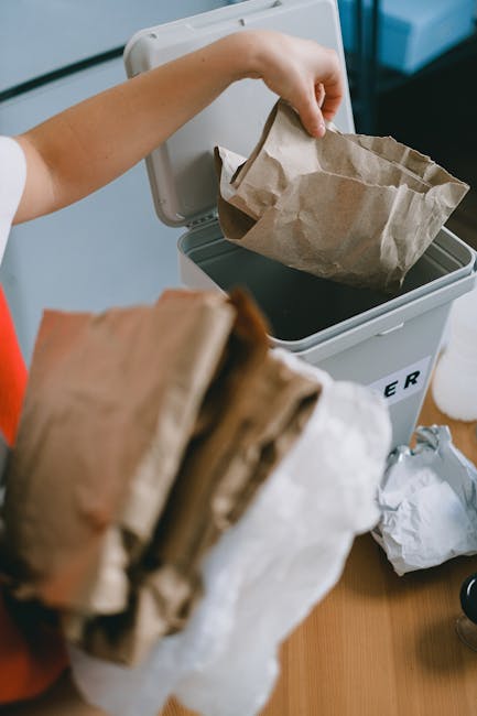 A person disposing of paper waste into a grey rubbish bin, with the lid open, inside a well-lit indoor space. The individual is holding a brown paper bag with crumpled edges, placing it into the bin, which appears to be designated for general waste collection. In the foreground, a stack of additional brown paper bags is visible, slightly blurred, indicating ongoing waste collection or removal activities. The background shows part of a countertop with a white object, possibly a plastic bottle or container, and the setting suggests an environment where waste is sorted and prepared for disposal or collection, aligning with private rubbish removal practices offered by Waste Collection Barnes. The scene emphasizes careful waste handling with an emphasis on paper waste, consistent with professional rubbish removal services for domestic or small-scale commercial purposes.