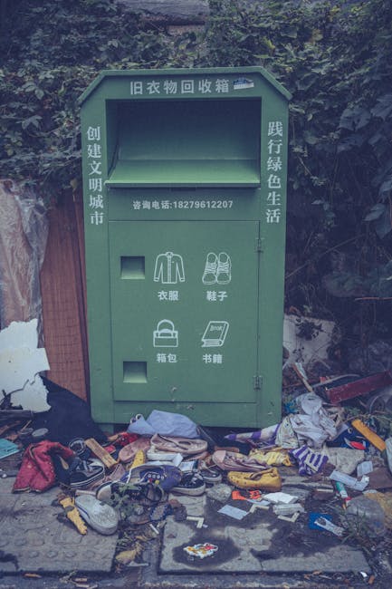 A green clothing donation collection box with Chinese characters and a contact phone number is positioned outdoors against a background of dense foliage and a wooden fence. The box appears to be made of metal with a slightly weathered finish, featuring several designated compartments for different types of clothing items: one for garments, with icons of shirts and jackets; another for shoes, depicted with shoe icons; a compartment for bags, illustrated with a handbag, and a section for books, represented by a stack of books. Surrounding the base of the container, there is a clutter of discarded items including shoes, clothing, plastic wrappers, and miscellaneous rubbish strewn across the ground, creating a messy scene in an outdoor setting. The scene suggests a misused collection point, with waste pile-up in front, and the environment around being somewhat unkempt. This visual context relates to the theme of rubbish removal, highlighting either illegal dumping or improper waste disposal at a public or charity collection point managed by waste collection services like Waste Collection Barnes, which specializes in alternative waste handling solutions.