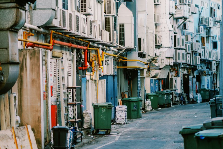 A narrow urban alleyway lined with a dense collection of air conditioning units and ventilation ducts mounted on the façades of multi-storey buildings. The wall surfaces are a mix of concrete and brick, with some areas showing signs of weathering and dirt accumulation. Numerous air conditioning units with white or grey casings, some with visible vents and grilles, are stacked unevenly, creating a textured, cluttered appearance. Bright red and yellow pipes run horizontally across the wall, connecting various equipment, and are supported by metal brackets. Several green plastic rubbish bins are placed along the pavement, some with white plastic bags or smaller containers beside them, indicating waste collection or ongoing clearance activities. A small black metal shelving unit and a few scattered bags or other items suggest a temporary storage or disposal zone. The lighting appears natural, casting soft shadows, emphasizing the utilitarian environment. The scene reflects an environment where private or building-specific waste handling, possibly by services like Waste Collection Barnes, might be necessary for alternative waste removal in confined or difficult-to-access areas, typical of urban alley clearing tasks.